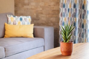 Uma planta em vaso sobre uma mesa de madeira, com um sofá cinzento e cortinas estampadas ao fundo.