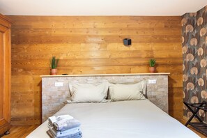 Quarto com parede de madeira, cama com duas almofadas, uma pequena planta no cabeceira e uma toalha dobrada no chão.