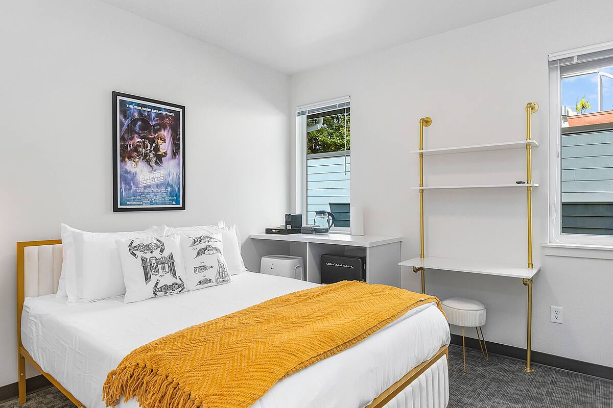 A hotel room with a bed, a desk, a chair, a shelf, and a window.