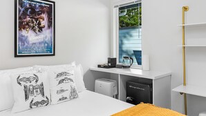 Una habitación de hotel con una cama, una ventana con persianas, un cartel de película enmarcado, una cafetera y un refrigerador negro.