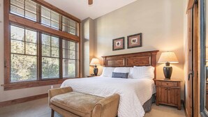 A bedroom with a large bed, two bedside lamps, a wooden bench, and a window with blinds. - Catamount Lodge 103 Two Bedroom (Truckee)