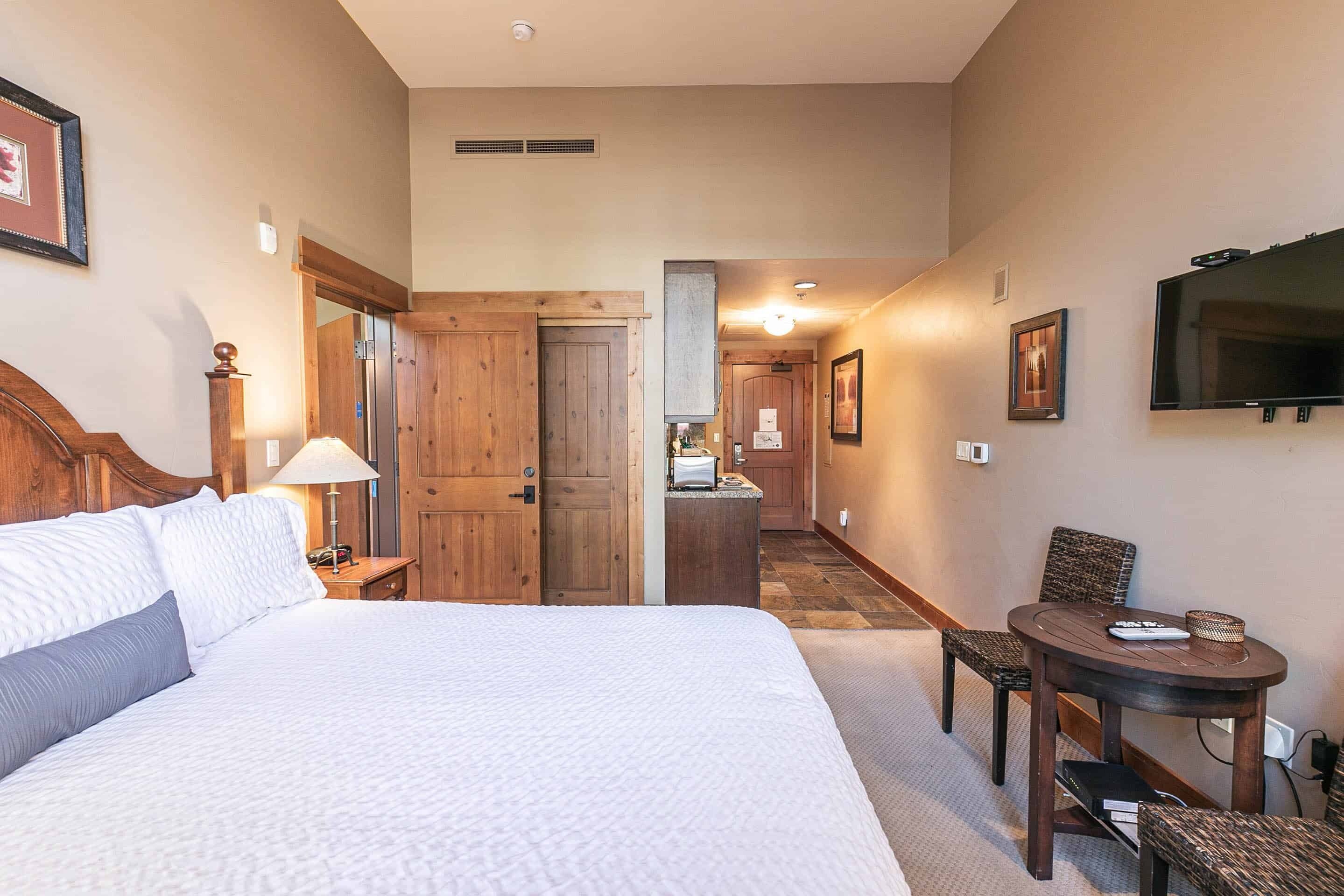 A hotel room with a large bed, a desk with a chair, a television, and a kitchenette in the background.