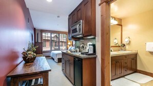 Cocina compacta con armarios de madera, microondas y lavavajillas. Hay una mesa de comedor con una planta, y se ve una cama al fondo.