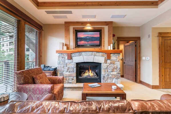 A cozy living room with a stone fireplace, leather sofa, and a television mounted above it. - Catamount Lodge 201 - 2 Bedroom Village Residence (Truckee)
