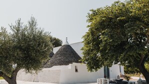 Een wit gebouw met een puntdak, omringd door olijfbomen en een geplaveid terras met zitplaatsen.