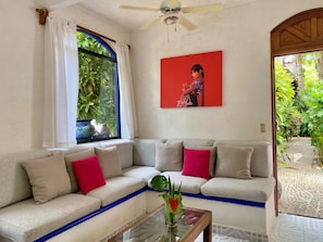 A living room with a beige sofa, red cushions, a glass coffee table, a ceiling fan, a window with white curtains, and a red painting. - Villa Tortolas 5 Min Walk To La Ropa Beach (Zihuatanejo)