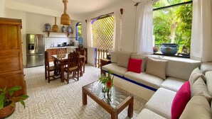 A living room with a sofa, a coffee table, and a dining area with chairs and a table. There is a kitchen in the background with a refrigerator and a water dispenser. - Villa Tortolas 5 Min Walk To La Ropa Beach (Zihuatanejo)
