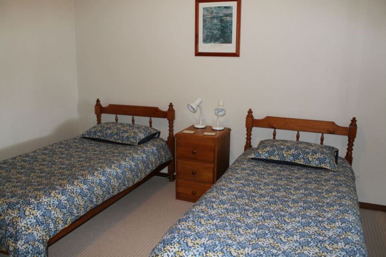 Una habitación con dos camas de madera, cada una con una colcha estampada y almohadas a juego. Hay una mesita de noche de madera con dos lámparas y un cajón pequeño. Encima de las camas, hay un cuadro enmarcado en la pared.
