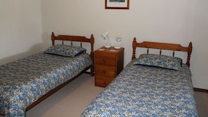 A room with two wooden beds, each with a floral bedspread and matching pillows. There is a wooden nightstand with two lamps and a small drawer. A framed picture hangs on the wall above the beds.