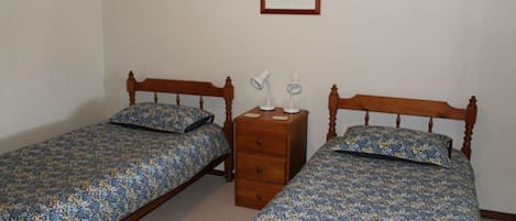 A room with two wooden beds, each with a floral bedspread and matching pillows. There is a wooden nightstand with two lamps and a small drawer. A framed picture hangs on the wall above the beds.