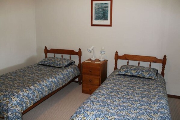 A room with two wooden beds, each with a floral bedspread and matching pillows. There is a wooden nightstand with two lamps and a small drawer. A framed picture hangs on the wall above the beds.