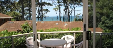 A balcony with two white plastic chairs and a small table, overlooking a coastal area with trees and buildings.