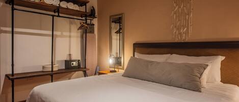 A neatly made bed with white linens, a bedside table with a lamp, and a shelf with folded towels and a radio.