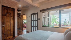 Une chambre à coucher avec un lit, une porte en bois, une fenêtre donnant sur des plantes et un ventilateur de plafond.