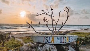 Un panneau sur lequel on peut lire « ISLA MUJERES », placé sur une côte rocheuse, avec vue sur l’océan et un coucher de soleil en arrière-plan.