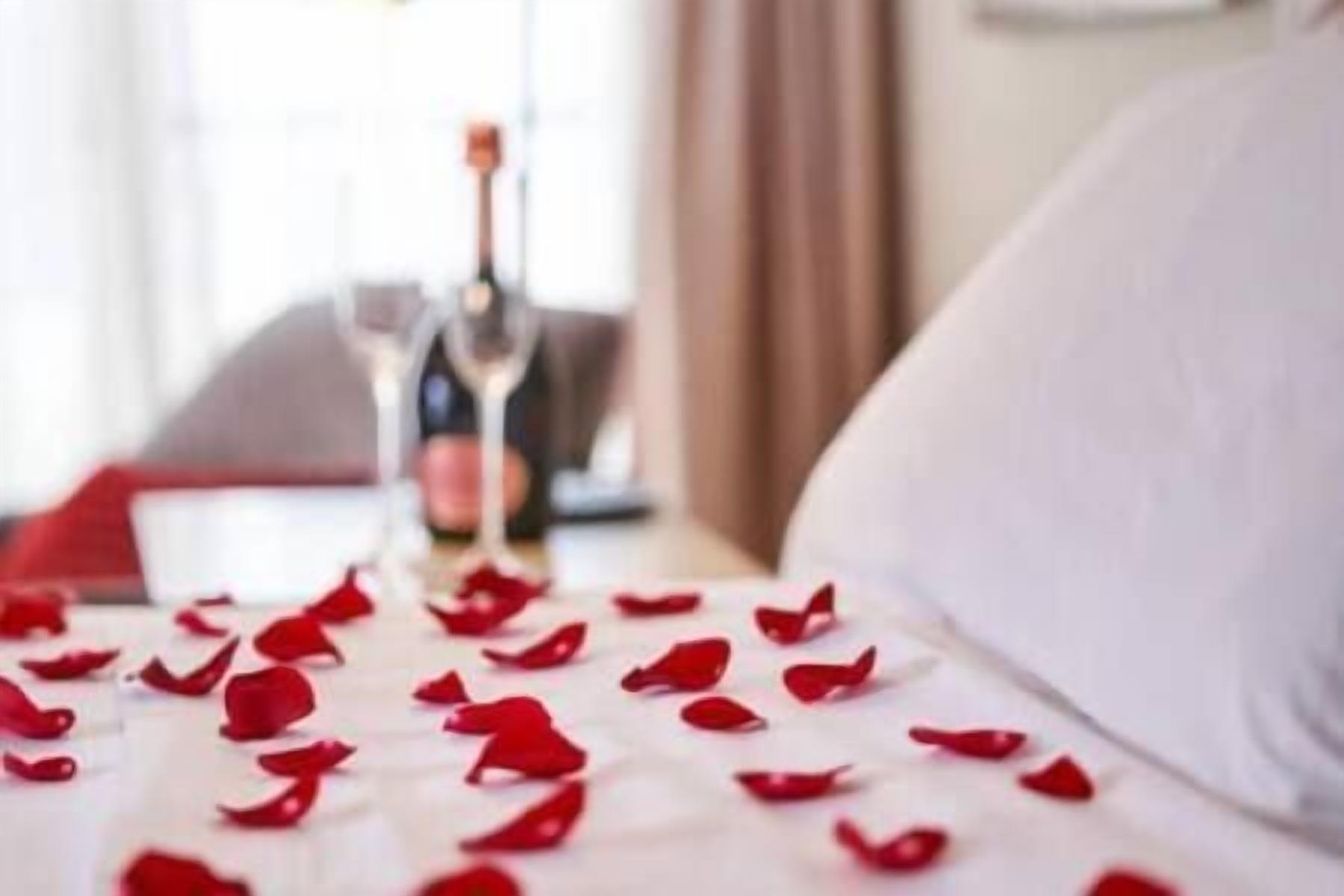 A bed with rose petals and a bottle of champagne in the background.