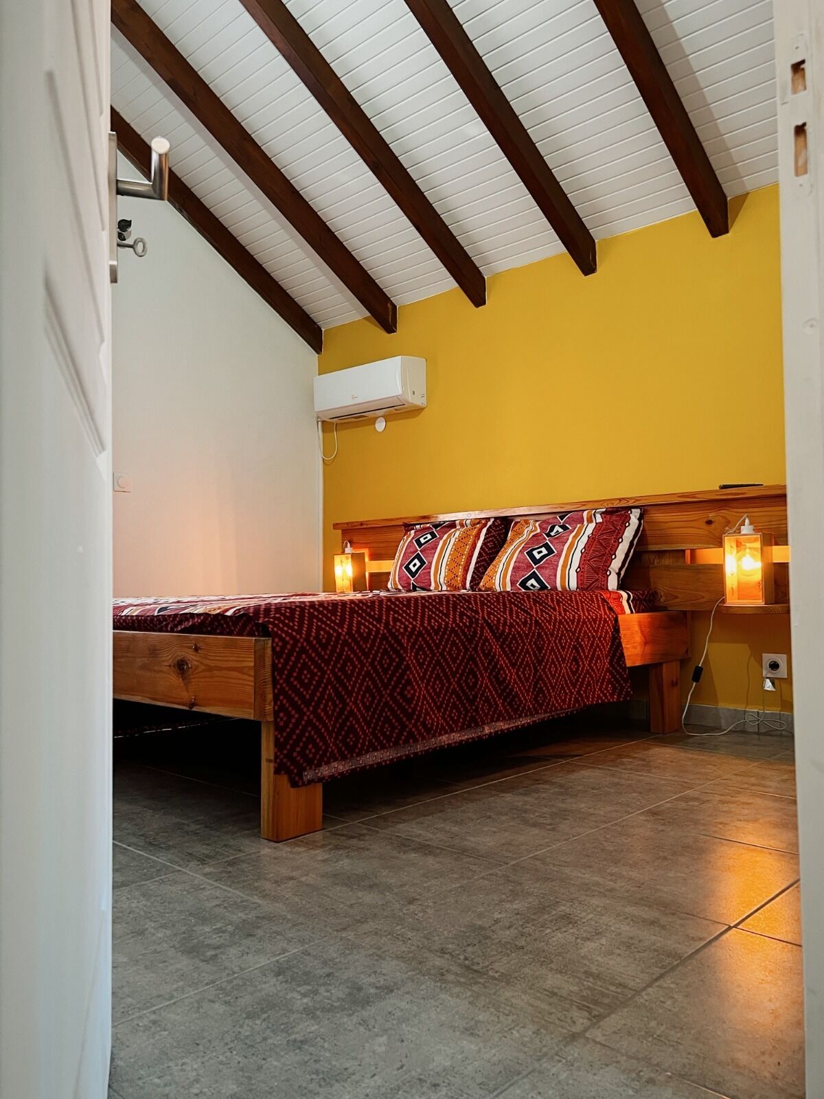 Une chambre avec un lit en bois, un mur jaune et un plafond en pente avec des poutres en bois.