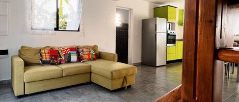 A modern living room with a sofa, a wooden bench, and a kitchen area with a refrigerator and cabinets.
