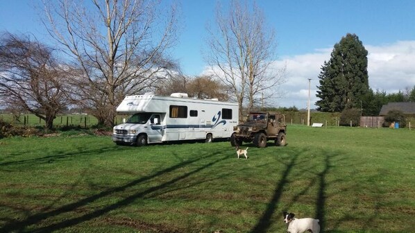 Una autocaravana y un vehículo militar estacionados en un campo cubierto de césped, con árboles y cielo despejado.