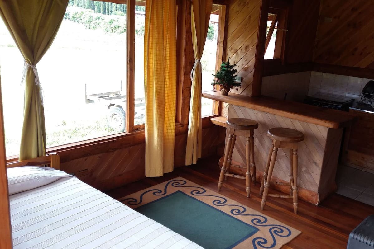A cozy cabin-style room with a bed, a small kitchen area with a bar stool, and a window with curtains.