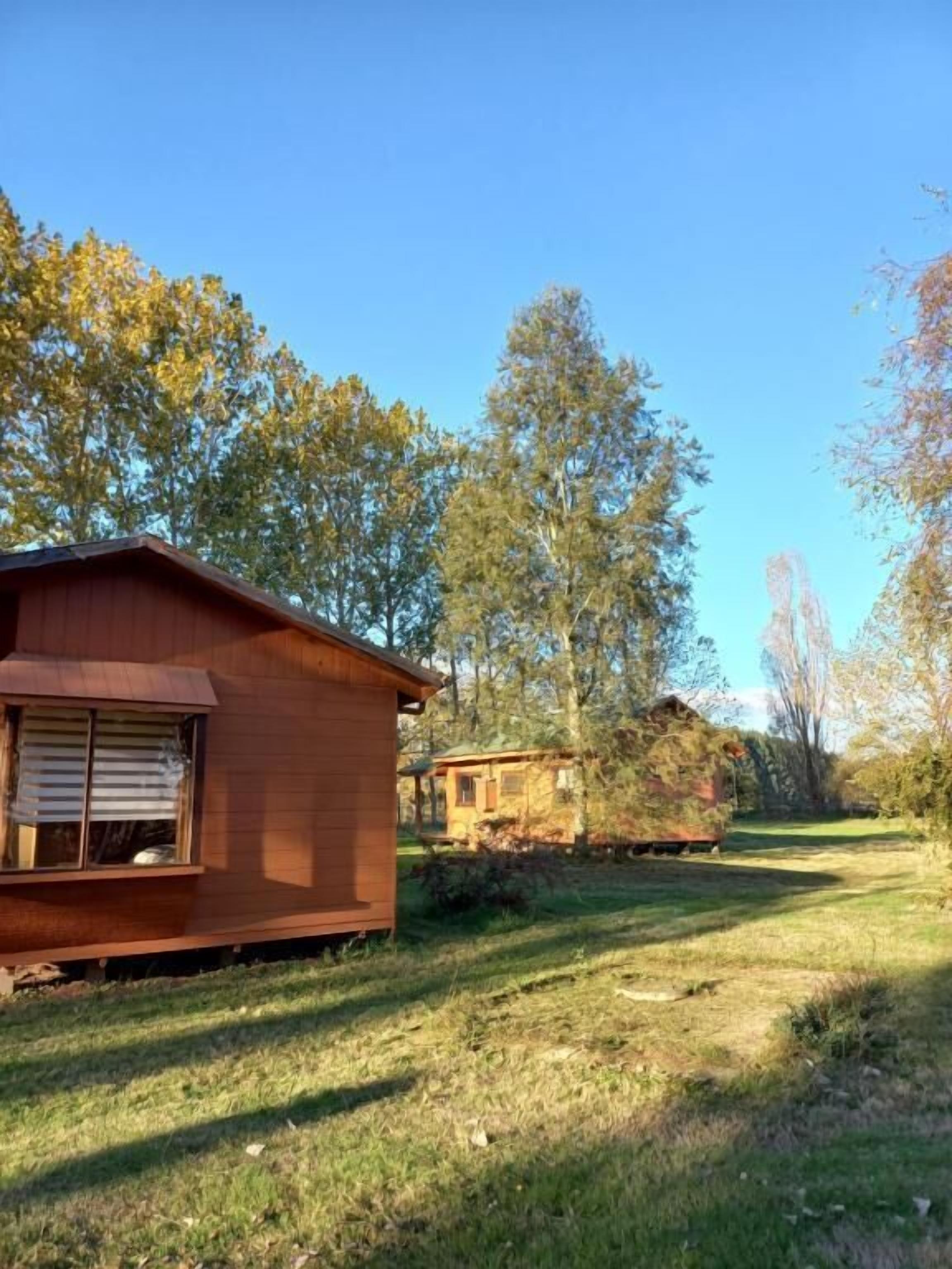 A rustic wooden cabin with a small attached shed, surrounded by a grassy area and trees.