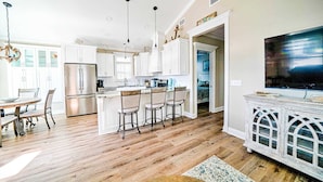 A spacious kitchen with white cabinets, a large island with bar stools, a stainless steel refrigerator, and a dining area with a wooden table and chairs.