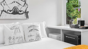 A bedroom with Star Wars-themed pillows, a window with a view of greenery, and a compact refrigerator. - Luke's Jedi Meditation Chamber (Seattle)