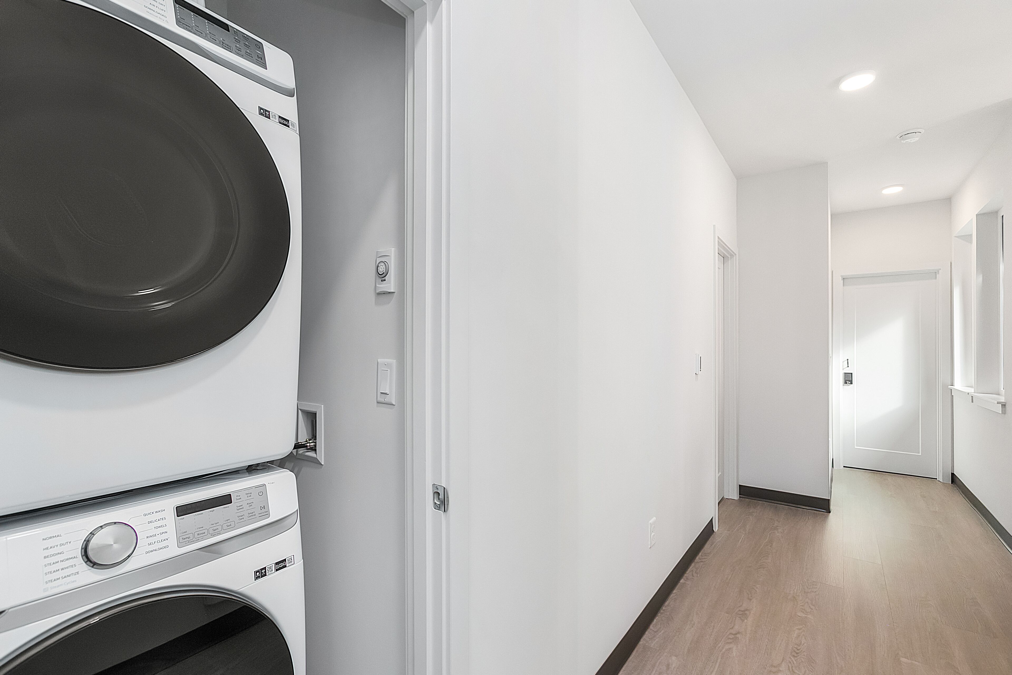 A laundry room with stacked washers and dryers, a white wall, and a wooden floor.