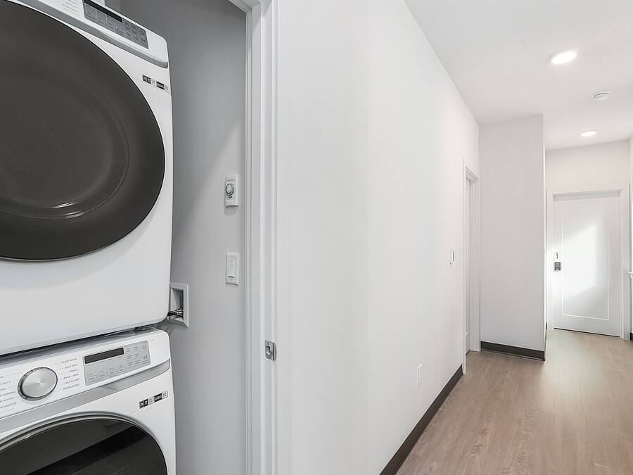 A laundry room with stacked washers and dryers, a white wall, and a wooden floor.
