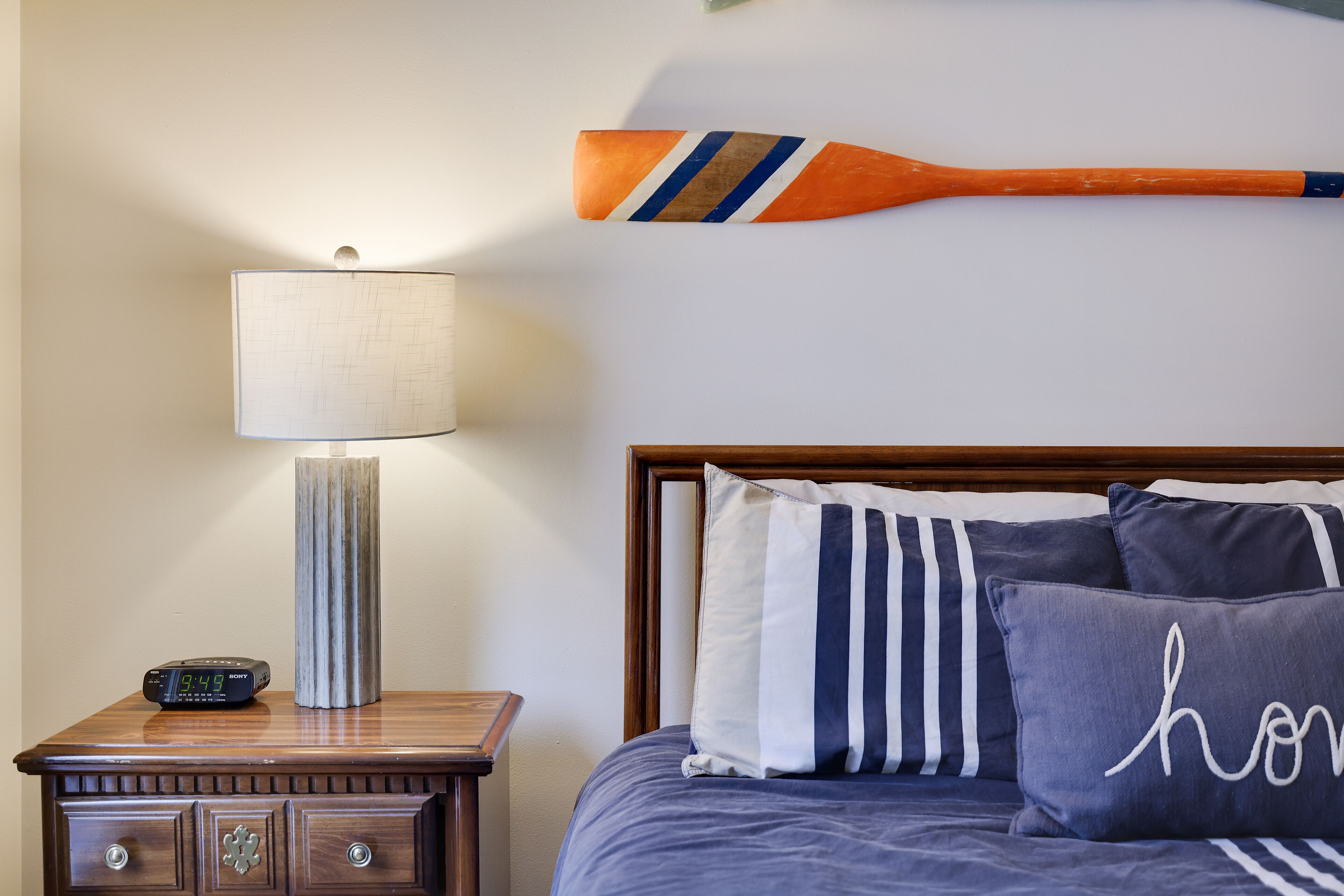 Ein ordentlich bezogenes Bett mit gestreiften Kissen, ein Nachttisch aus Holz mit einer Lampe und ein an der Wand befestigtes Paddel.