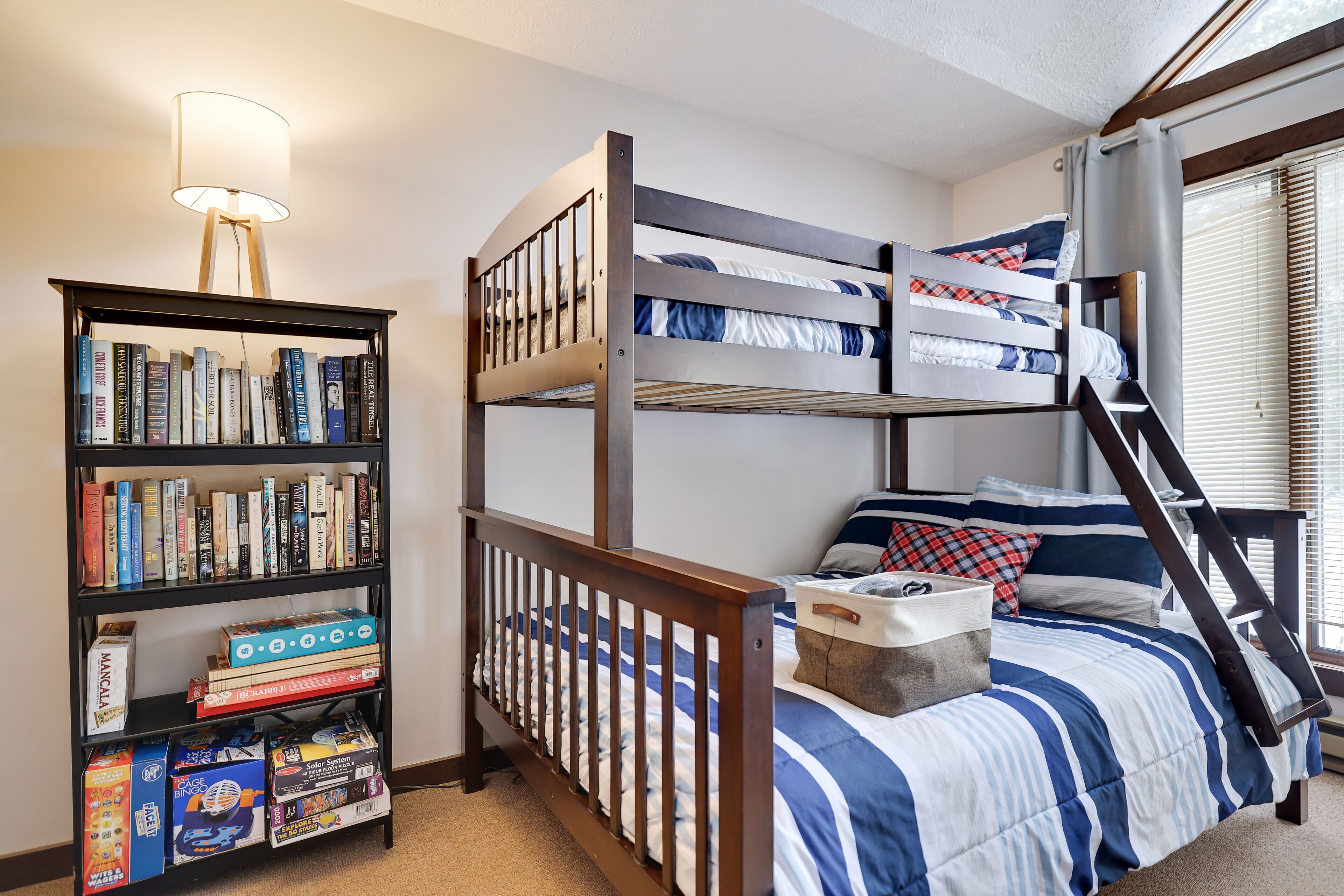 A bunk bed with a striped bedspread, a bookshelf with various books and boxes, and a lamp.