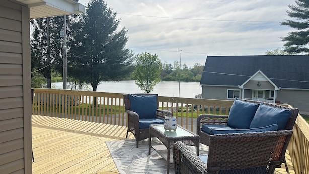 En overdækket terrasse med fletmøbler og et glasbord, med udsigt over en sø og et hus.