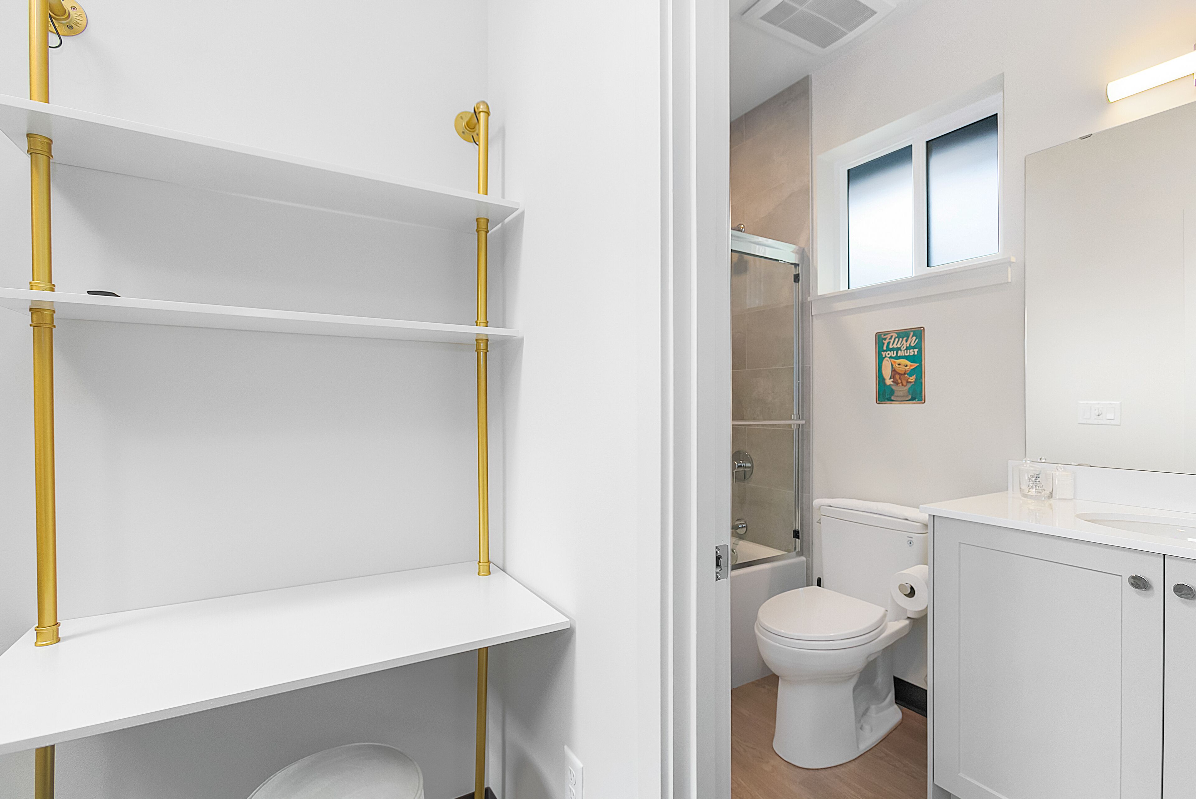A modern bathroom with white fixtures, a toilet, a sink, and a shower. There are gold-colored pipes and a shelf unit.