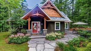 Un chalet rustique avec un sentier de pierres menant à une porte en bois, entouré d’une végétation luxuriante et doté d’un patio en pierre avec des sièges d’extérieur.