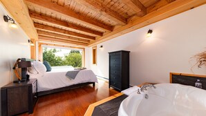 Una camera da letto con un letto grande, soffitto in legno, armadio nero, bagno con vasca e finestra vista alberi.
