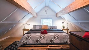 Une chambre avec un plafond en pente, des poutres en bois, un lit avec une literie à motifs, des tables de chevet avec des lampes et une fenêtre avec des stores.