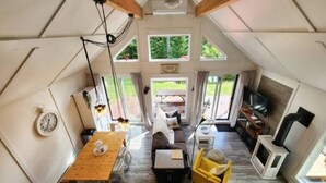 Un intérieur chaleureux et bien éclairé, avec une table à manger en bois, un canapé et un fauteuil jaune. On aperçoit une cheminée sur la droite, et de grandes fenêtres laissent entrer la lumière naturelle.
