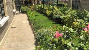 A paved pathway leading to a building with a red-tiled roof, surrounded by lush greenery and flowering plants. - Splendid 4-Bedroom with A Pool (Athi River)