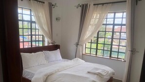 A neatly made bed in a room with large windows, curtains, and a view of a residential area. - Splendid 4-Bedroom with A Pool (Athi River)