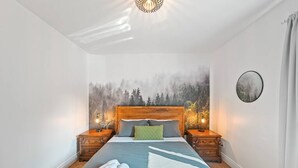Una cama bien tendida con una colcha azul, un almohadón verde y toallas enrolladas en el suelo. Hay dos mesitas de noche con lámparas, un cuadro grande de un bosque en la pared y un espejo redondo en la pared.