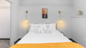 A neatly arranged hotel room with a bed, two bedside lamps, a poster on the wall, and a small table with a lamp. - Skywalker Rebel Base (Seattle)