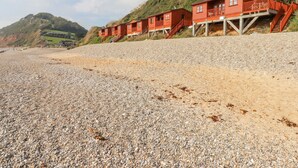 Un hotel costero con edificios de madera roja, una playa de guijarros y un paisaje montañoso al fondo.