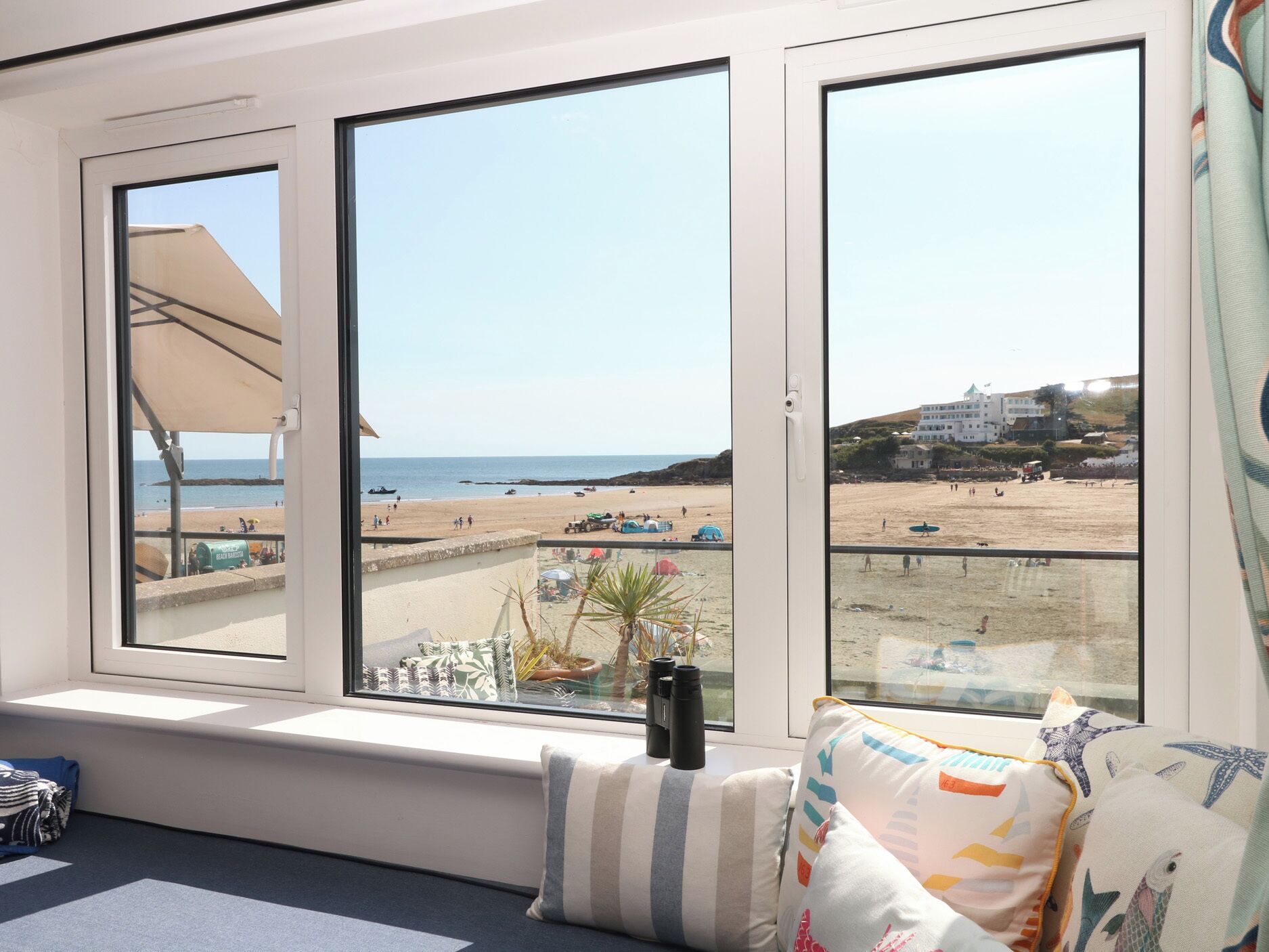 A room with a large window overlooking a beach, a bed with striped pillows, and a pair of binoculars on the windowsill.
