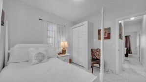 A white bedroom with a bed, nightstand, lamp, chair, and a wardrobe.