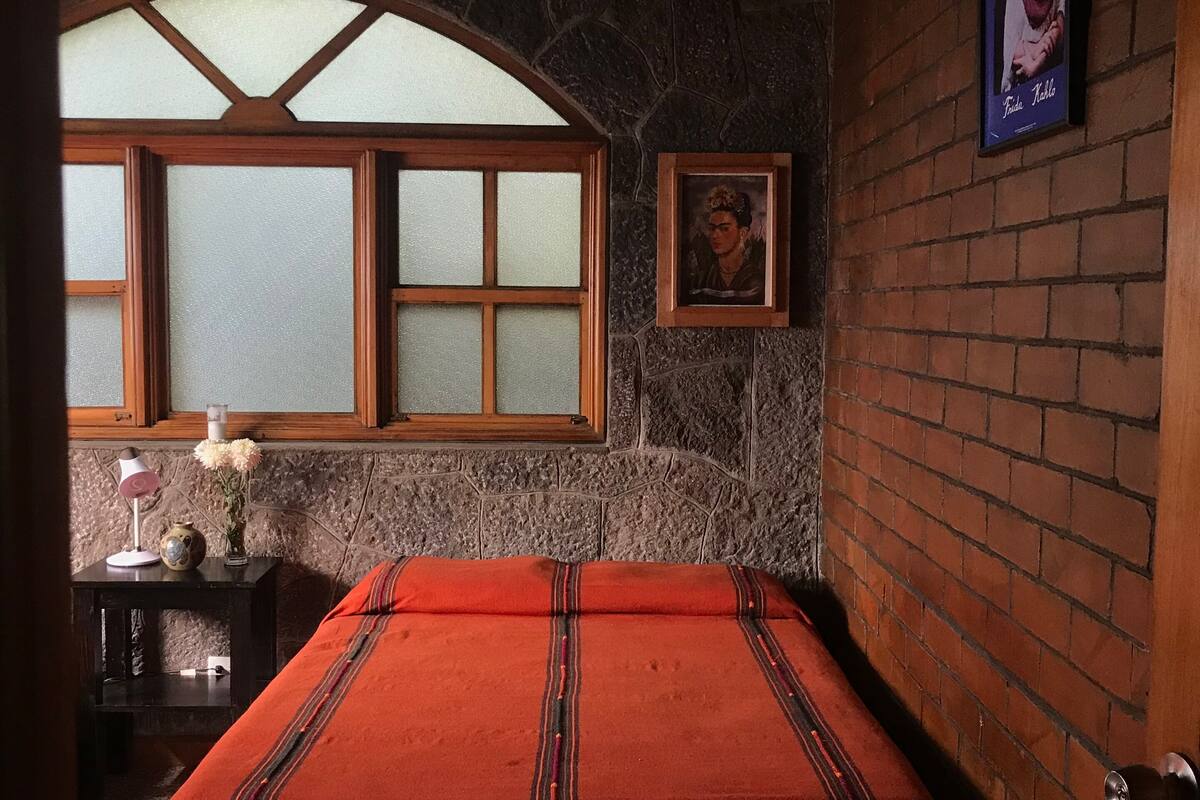Un dormitorio con pared de piedra, ventana con marco de madera, cama con manta roja, mesita de noche con lámpara y un cuadro enmarcado en la pared.