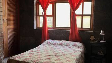 Ein Schlafzimmer mit Bett, einem Fenster mit Vorhängen, einem Nachttisch und einer Wand mit strukturierter Oberfläche.