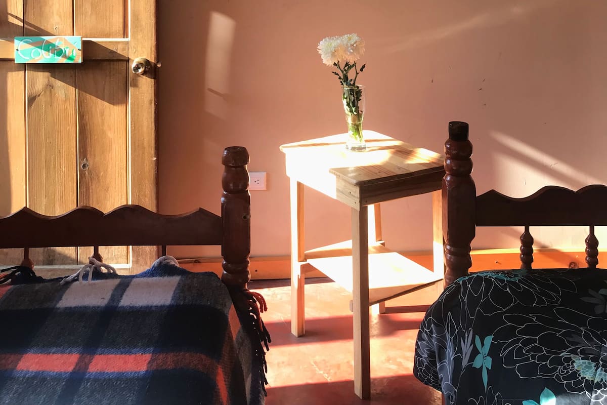Una habitación con una puerta de madera, una mesita con un jarrón de flores y una cama con una colcha escocesa.