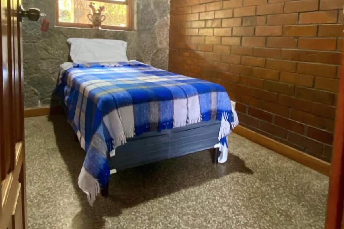 Un dormitorio con pared de piedra, pared de ladrillo, ventana con detalle decorativo y cama con colcha azul y blanca a cuadros.