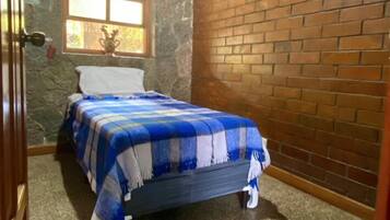 Un dormitorio con pared de piedra, pared de ladrillo, ventana con detalle decorativo y cama con colcha azul y blanca a cuadros.