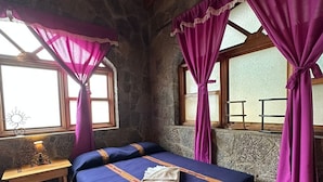 A stone-walled room with a bed, a wooden table, and purple curtains. - Casa Qatzij - Guest House (San Lucas Toliman)